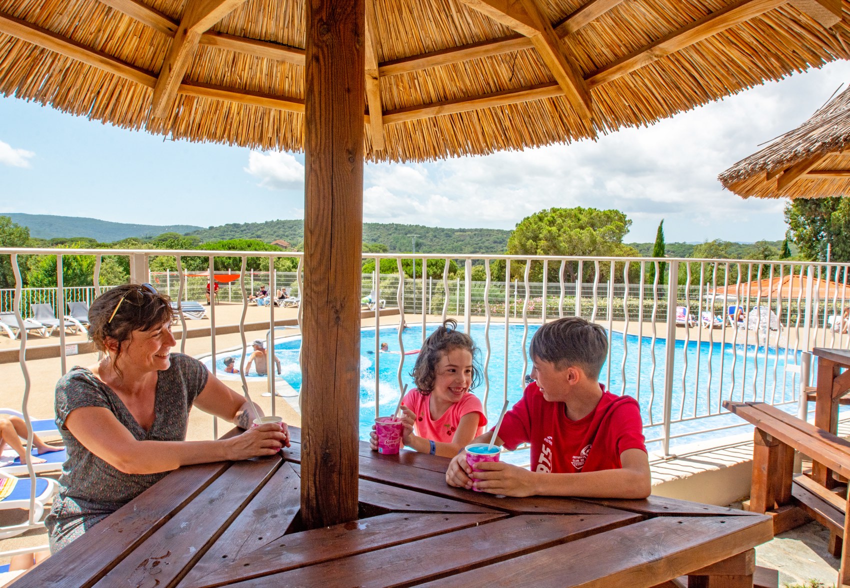 Eine Familie sitzt an einem Tisch unter dem Strohdach mit Blick auf den Swimmingpool